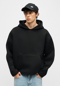 Νεαρός άνδρας που φοράει μαύρο φούτερ με κουκούλα και μαύρο καπέλο Nike, με τα χέρια στις τσέπες ανοιχτόχρωμων μπλε τζιν, κοιτάζοντας προς το πλάι μπροστά σε έναν απλό φόντο.