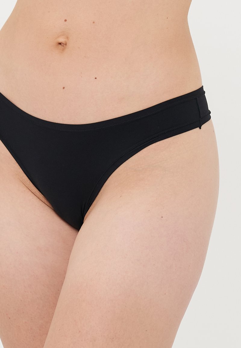 Primer plano de una persona que lleva ropa interior básica tipo bikini negra, mostrando el abdomen inferior y la parte superior de los muslos sobre un fondo neutro.