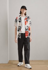 Camisa de manga corta con un patrón gráfico audaz en rojo, negro y beige, combinada con pantalones negros y zapatillas blancas.