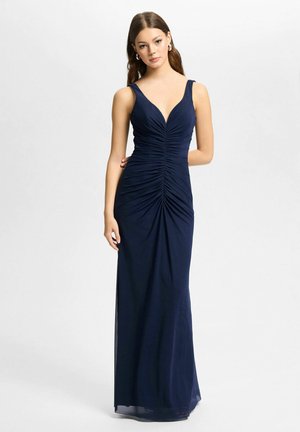 Woman wearing a navy blue sleeveless ruched gown with a deep V-neck, standing against a plain light background.