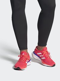 Zapatillas deportivas de un color rosa brillante con rayas blancas y detalles en púrpura. Parte superior de malla con texturas, combinadas con leggings negros.