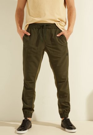 Mann trägt olivgrüne Jogginghose mit elastischen Bündchen und schwarzen Sneakers, Hände in den Taschen, beigefarbenes Hemd teilweise eingesteckt.