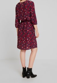 Plynoucí burgundská šaty s tečkovaným vzorem v červené, růžové a modré. Tříčtvrteční rukávy, zúžený pas a detaily ve tvaru klíčové díry na zádech. Černé kotníkové boty.