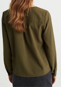 Olivgrön långärmad blus med strukturfinish, rund halsringning, ryggsöm och knappade manschetter, matchad med svarta byxor.