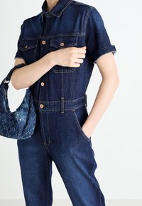 Combinaison en denim bleu foncé à manches courtes, avec deux poches poitrine et des boutons dorés. Associée à un sac à main bleu texturé.