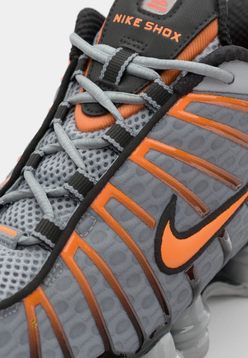Les chaussures Nike Shox présentent une tige en mesh gris avec des accents orange, un système de laçage distinct et un design texturé. La semelle comprend un amorti visible.