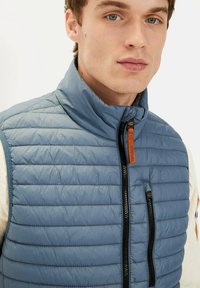 Hellblaues Pufferwesten mit hohem Kragen, mit horizontalen Steppnähten, zwei Reißverschlusstaschen und einem Reißverschlussgriff aus braunem Leder.