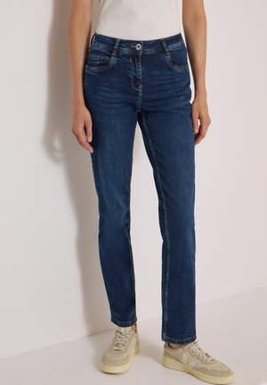 Person iført mørkeblå jeans med lige ben og beige sneakers, der står mod en lys baggrund.