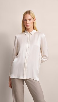 Cream-colored, long-sleeve blouse made of smooth fabric with a button-up front, pointed collar, and subtle sheen, paired with light beige trousers.