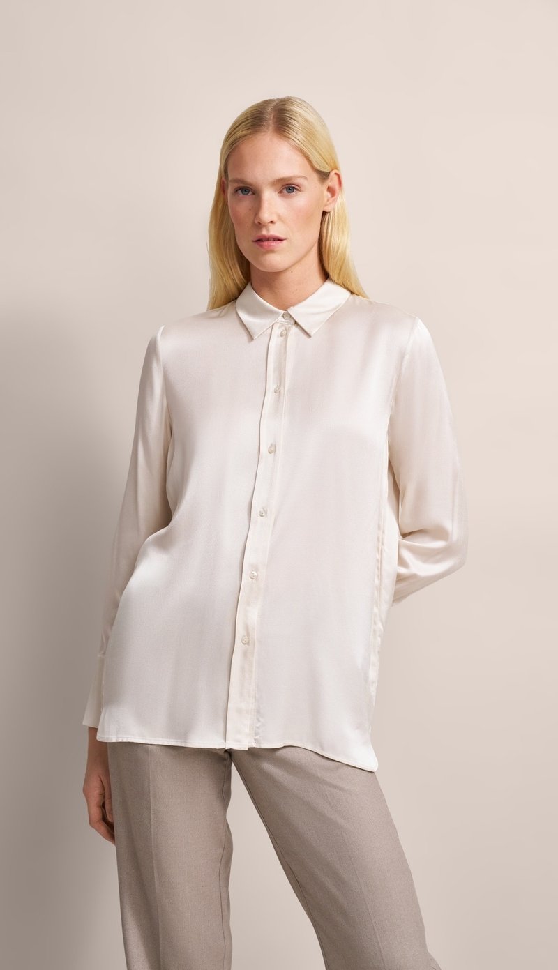 Cream-colored, long-sleeve blouse made of smooth fabric with a button-up front, pointed collar, and subtle sheen, paired with light beige trousers.