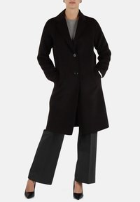 Cappotto di lana nero con revers a punta, chiusura con un bottone e due tasche laterali. Abbinato a pantaloni grigi a gamba larga e tacchi neri.