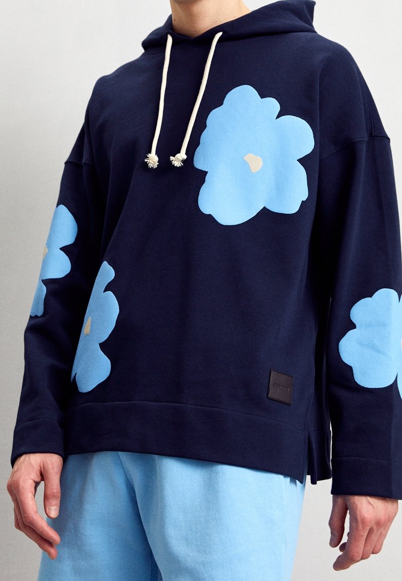 Felpa con cappuccio blu navy con grandi motivi a fiori azzurri, cordini bianchi e etichetta in pelle marrone. Abbinata a pantaloni azzurri.