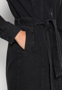 Musta farkkukotime, jossa on vyötärönauha, nappikiinnitys, pitkät hihat ja sivutaskut. Kankaan pinta on teksturoitu ja siinä on hieman kulunut ilme.