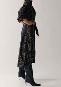 Longue robe marine avec motif de chaînes dorées, taille cintrée et manches bouffantes. Portée avec des bottes noires à talons hautes jusqu'aux genoux. Texture de tissu lisse.