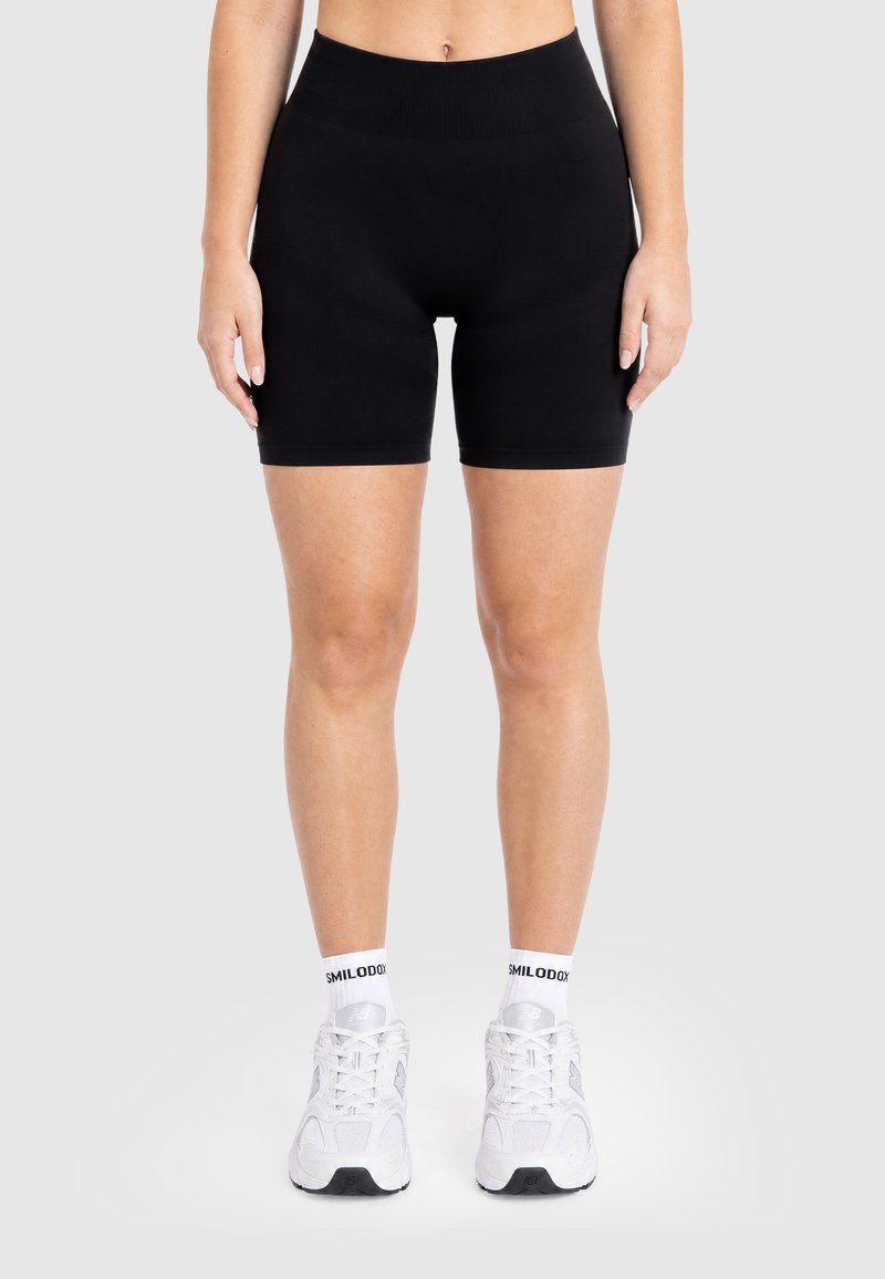 Person stående iført sorte tætsiddende shorts med høj talje, hvide sokker med tekst og hvide sportssko mod en ensartet baggrund.