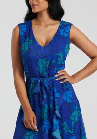 Blå blommor klänning med V-ringning, ärmlös design och knytbälte. Har blå och turkosa mönster på ett lätt tyg med en flödande silhuett.