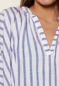 Blusa bianca con strisce blu, colletto e dettagli a plissettatura. Realizzata in tessuto leggero, presenta fantasie verticali e orizzontali.