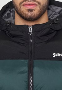 Svart och grön pufferjacka med dragkedja, justerbar huva och förstärkt sömnad. Har en logotyp på bröstet och en slät yta.