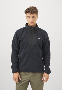 Sort fleece-pullover med høj krave, halv-lynlås foran og sidelommer med lynlås. Har en glat tekstur og et logo på brystet.