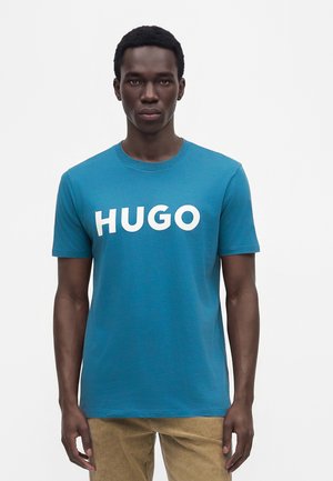Млад мъж, носещ синя тениска с бял "HUGO" текст, стоящ с неутрално изражение на лице пред обикновен бял фон.