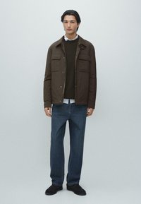 Brun jacka med dragkedja och fyra framfickor, bärs över en mörkgrön tröja och vit skjorta, kombinerad med blå jeans och svarta skor.