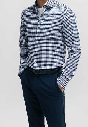 Man wearing navy blue and white checkered button-up shirt and navy blue pants with black belt, hands in pockets, against plain background.