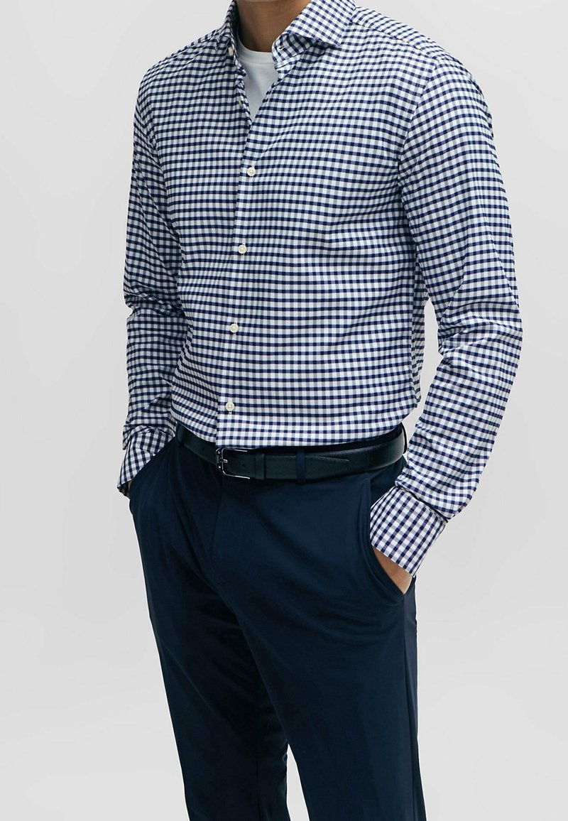 Homme portant une chemise à carreaux bleu marine et blanche à boutons et un pantalon bleu marine avec une ceinture noire, les mains dans les poches, sur un fond uni.