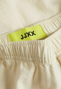 Tejido color crema con una cintura elástica, con una etiqueta verde brillante que tiene el texto "JJXX" en negro. Textura suave y diseño sencillo.