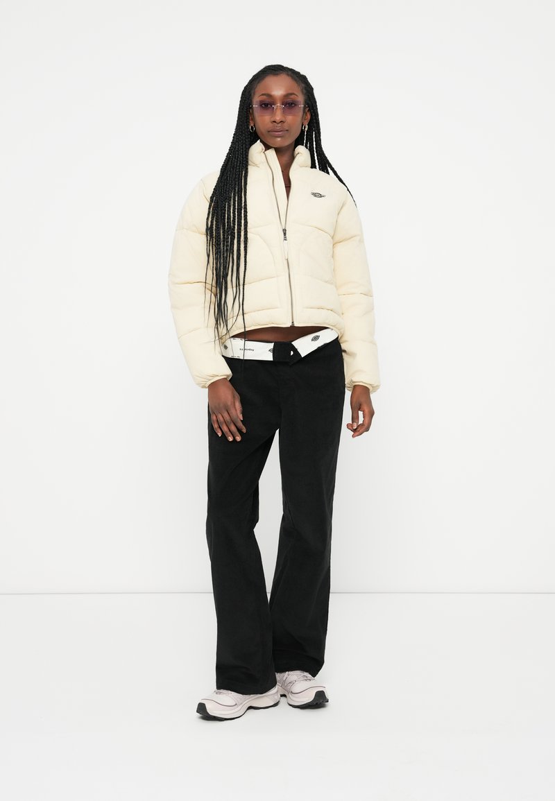 Doudoune crème avec un col montant et une fermeture éclair, pantalon en velours côtelé noir et baskets de couleur claire. Cheveux longs tressés et lunettes de soleil violettes.