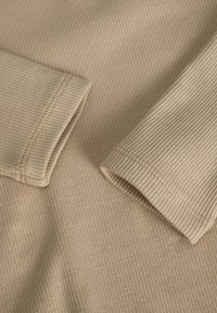 Beige bordázott anyag puha tapintással, gondosan kidolgozott mandzsettákkal és kontrasztos varrási részletekkel a széleken.