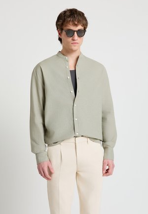Uomo con camicia verde chiaro colletto mandarino sopra una maglia nera, pantaloni crema e occhiali da sole scuri, in piedi davanti a uno sfondo semplice.