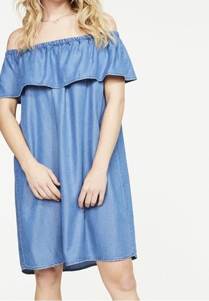 Femme portant une robe en denim bleue à épaules dénudées avec un détail à volants, debout les mains jointes devant un fond blanc.