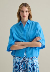 Camisa azul claro de manga corta y botón, con un corte relajado, bolsillo único en el pecho y una textura suave. Llevada sobre un vestido azul con estampado.