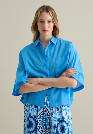 Camisa azul claro de manga corta y botón, con un corte relajado, bolsillo único en el pecho y una textura suave. Llevada sobre un vestido azul con estampado.