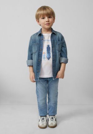 Jeanshemd over een wit graphic t-shirt, lichtblauwe spijkerbroek en sneakers. Korte blonde haren, staand met handen langs de zij.