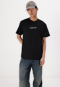T-shirt en coton noir à manches courtes avec un logo brodé blanc sur la poitrine, associé à un pantalon en denim bleu, offrant une coupe décontractée.