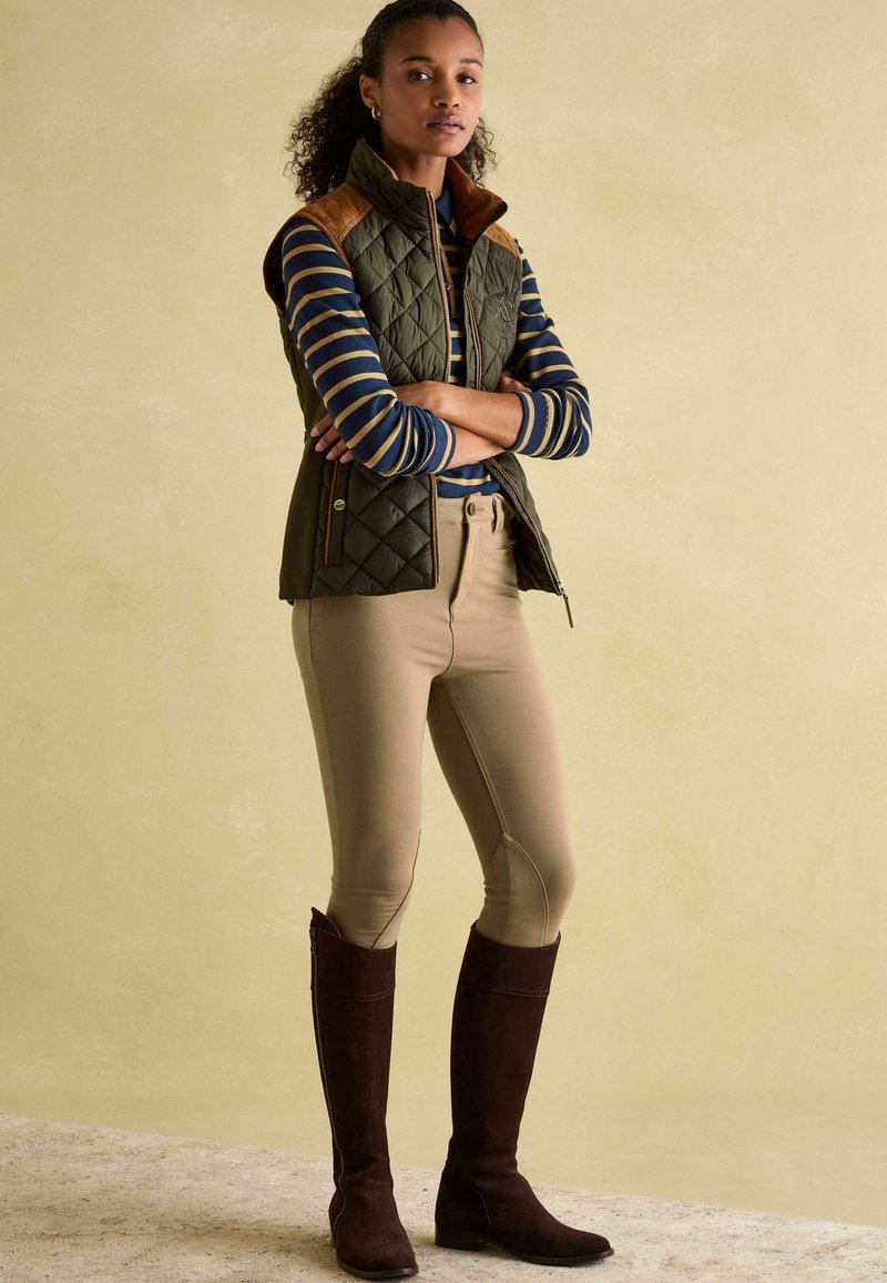 Prešívaná vestu v olivovej zelenej farbe s hnedými zvýrazneniami na pleciach, spárovaná s námornícky modrým a bielym pruhovaným tričkom, béžovými jazdeckými nohavicami a hnedými semišovými čižmami.