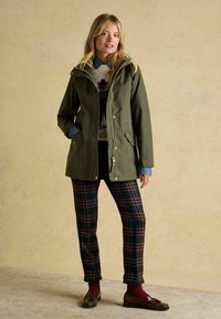 Zielona wodoodporna parka z kapturem, na zamek błyskawiczny i bocznymi kieszeniami. Nosi się ją na wzorzystym swetrze i w kratę spodniach, do tego brązowe mokasyny.