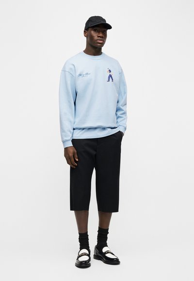 Άνδρας που φοράει ανοιχτό μπλε φούτερ με σχέδιο, μαύρο καπέλο, μαύρο κοντό παντελόνι, μαύρες κάλτσες και μαύρα-άσπρα loafers, στέκεται μπροστά σε λευκό φόντο.