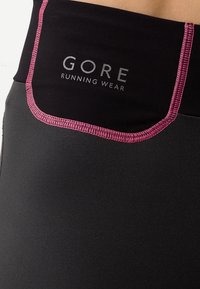 Černé běžecké šortky s páskem zvýrazněným v růžové barvě. Na boku mají stříbrně tištěné logo "GORE RUNNING WEAR". Hladký, elastický materiál.