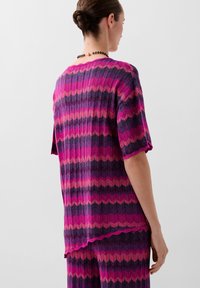 Knit top with ribbed texture in shades of pink and purple, featuring a zigzag pattern and scalloped hem. Short sleeves and relaxed fit.