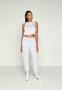 Mujer de pie vistiendo una parte superior sin mangas blanca y corta, pantalones jogger grises claros y zapatillas blancas con rayas negras sobre un fondo liso.