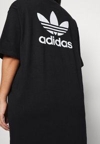T-shirt en coton noir avec une coupe oversize. Logo Adidas blanc et design trèfle au dos, avec trois rayures horizontales.