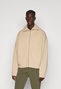 Giacca a zip beige a coste con colletto, caratterizzata da una vestibilità rilassata e maniche lunghe. Indossata con pantaloni verde oliva.