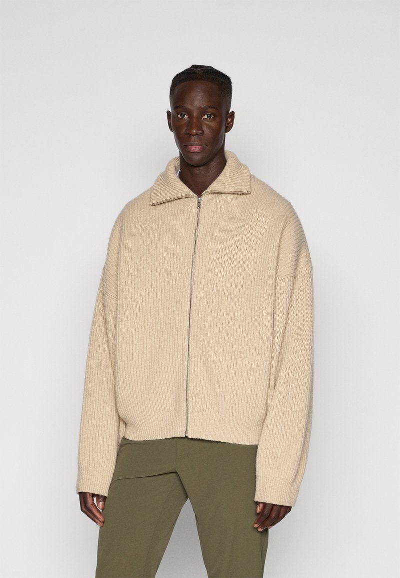 Giacca a zip beige a coste con colletto, caratterizzata da una vestibilità rilassata e maniche lunghe. Indossata con pantaloni verde oliva.
