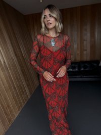 Μακρύ κόκκινο φόρεμα με διαφανές επικάλυμμα που φέρει μοτίβο μαύρου φύλλου και λεοπάρ. Τονίζεται από ένα μεγάλο κολιέ, εκτεθειμένο σε εσωτερικό χώρο.