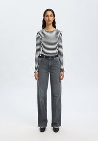 Camisa de manga longa às riscas em preto e branco, combinada com calças cinzentas de cintura alta e pernas largas, complementada por um cinto preto e sapatos pretos.