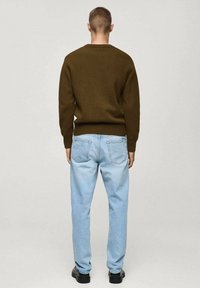 Brauner Strickpullover mit geripptem Saum, kombiniert mit hellblauen Jeans aus Denim, die über Gesäßtaschen und einen lässigen Schnitt verfügen. Schwarze Schuhe runden das Outfit ab.