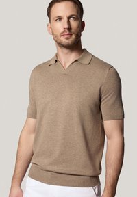 Beżowa koszulka polo z krótkim rękawem, z dekoltem w serek i ściągaczem na dole. Miękka faktura i czysty design. Noszona z białymi spodniami.