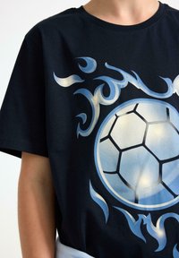 Sort bomulds T-shirt med et stort blåt og sølvfodboldgrafik omgivet af flamelignende detaljer i nuancer af blå.
